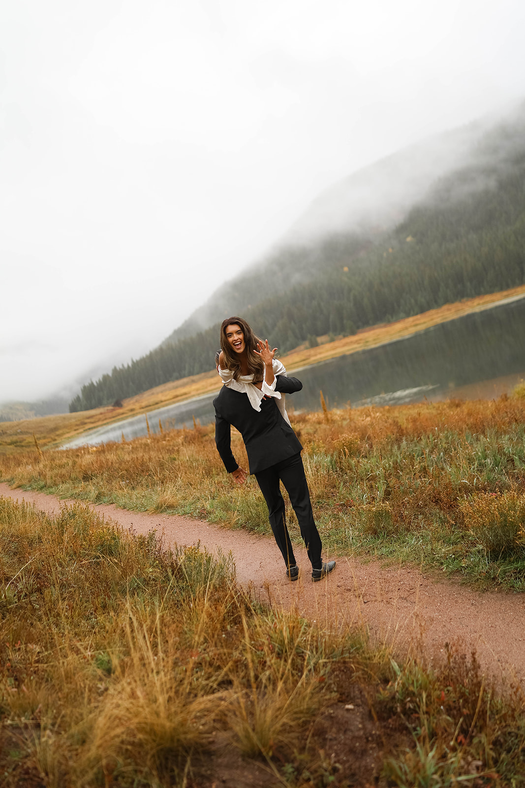 Bride laughing while being carried piggyback by the groom along a dirt path near a lake, with misty mountains and fall grass surrounding them.