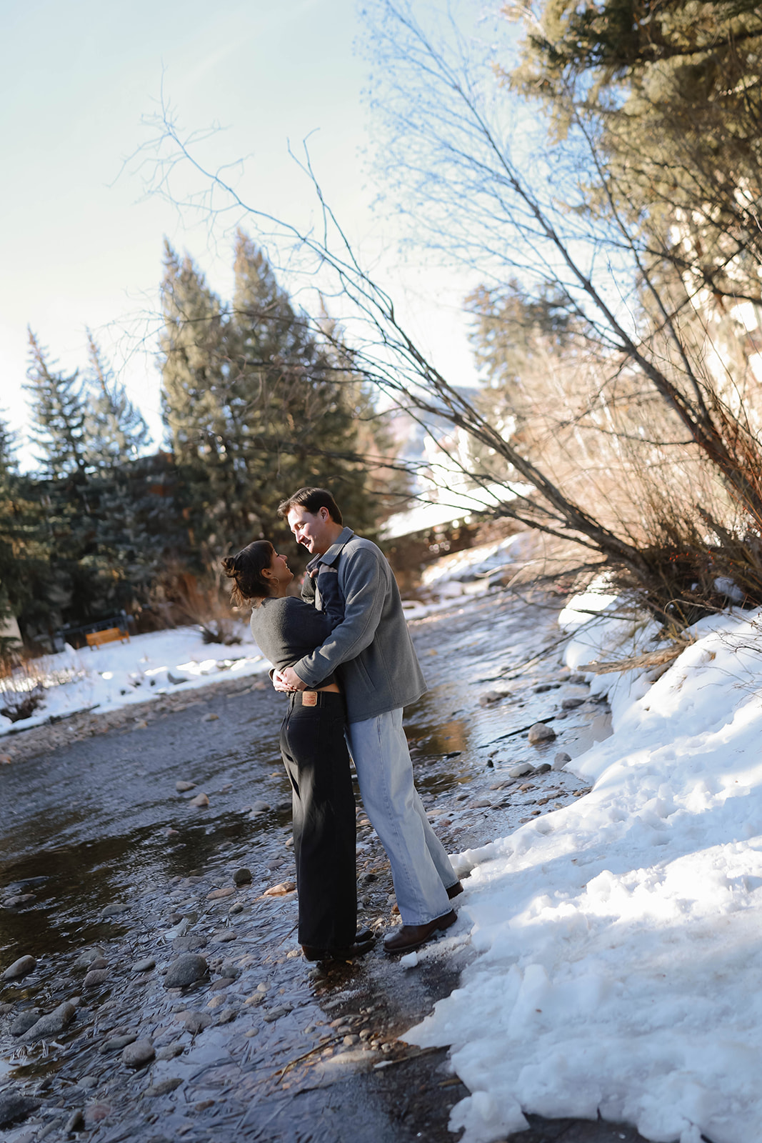 Couple embracing beside a snowy creek, wrapped in winter layers, with evergreen trees and fresh snow creating a peaceful alpine setting.