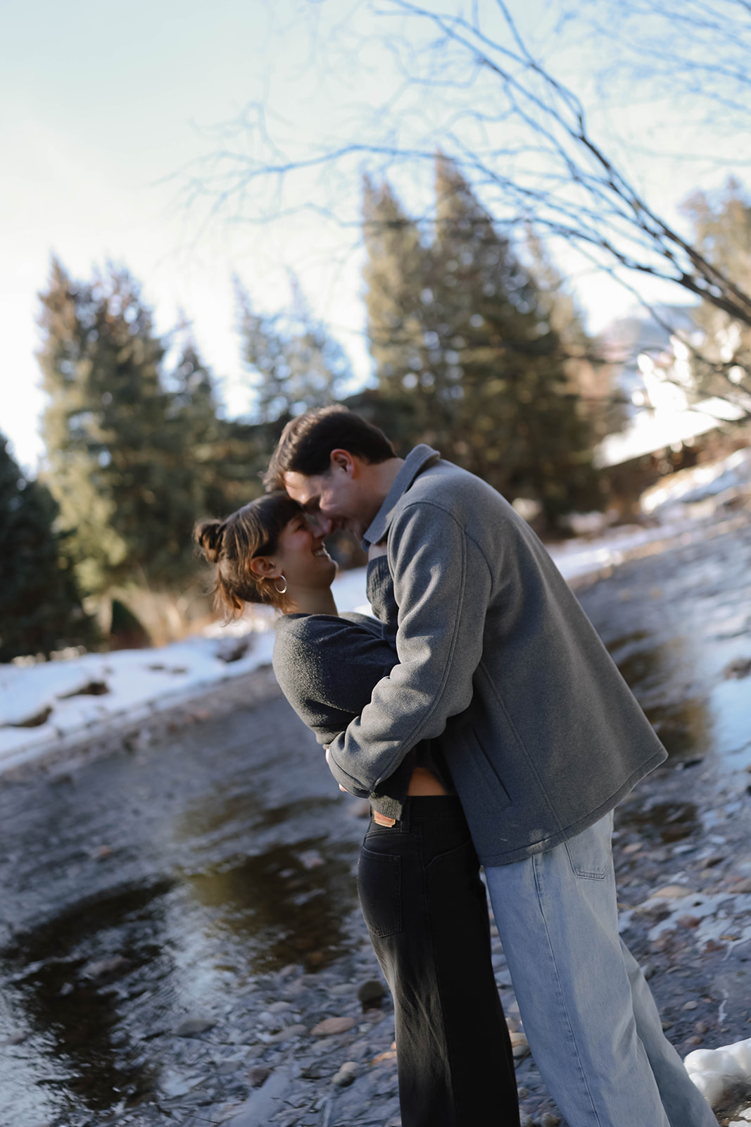 A couple standing close together along a shallow creek, wrapped in each other’s arms with snow, trees, and mountain scenery in the background.