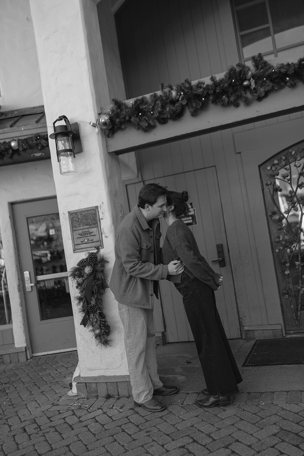 Black-and-white photo of a couple leaning in for a kiss outside a charming mountain building decorated with holiday garland.