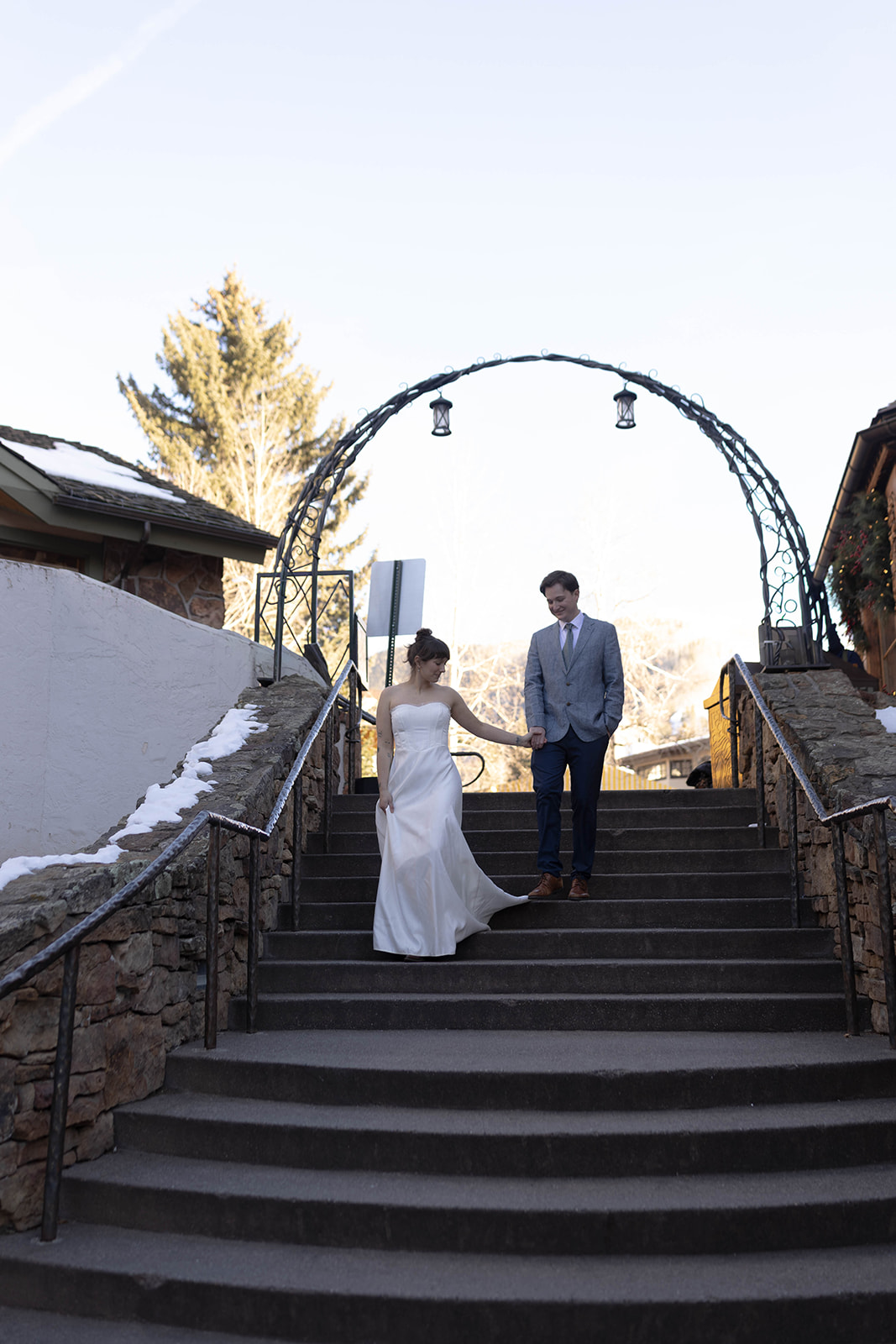 Bride and groom walking hand in hand down stone steps beneath an iron archway during a winter vail elopement in the village.