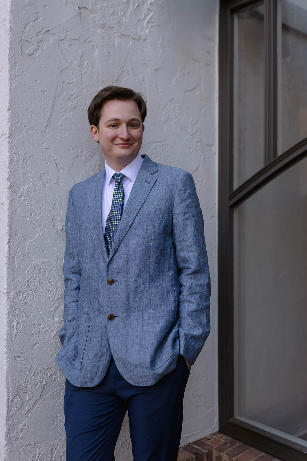 Groom standing casually in a blue blazer and tie against a light stucco wall for a relaxed wedding portrait.