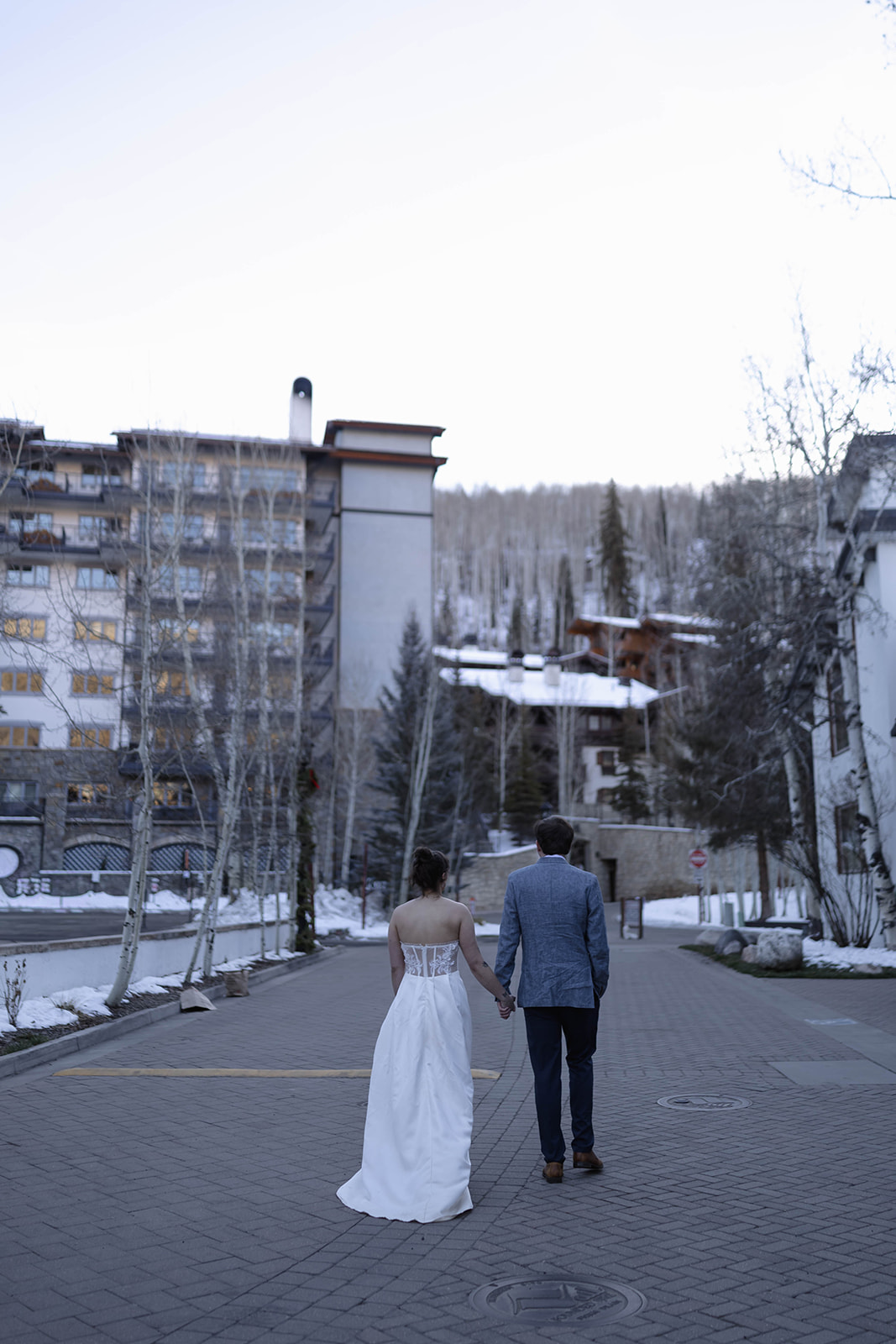 Couple walking away hand in hand through a quiet mountain village street, framed by winter trees and alpine architecture after their elopement ceremony.