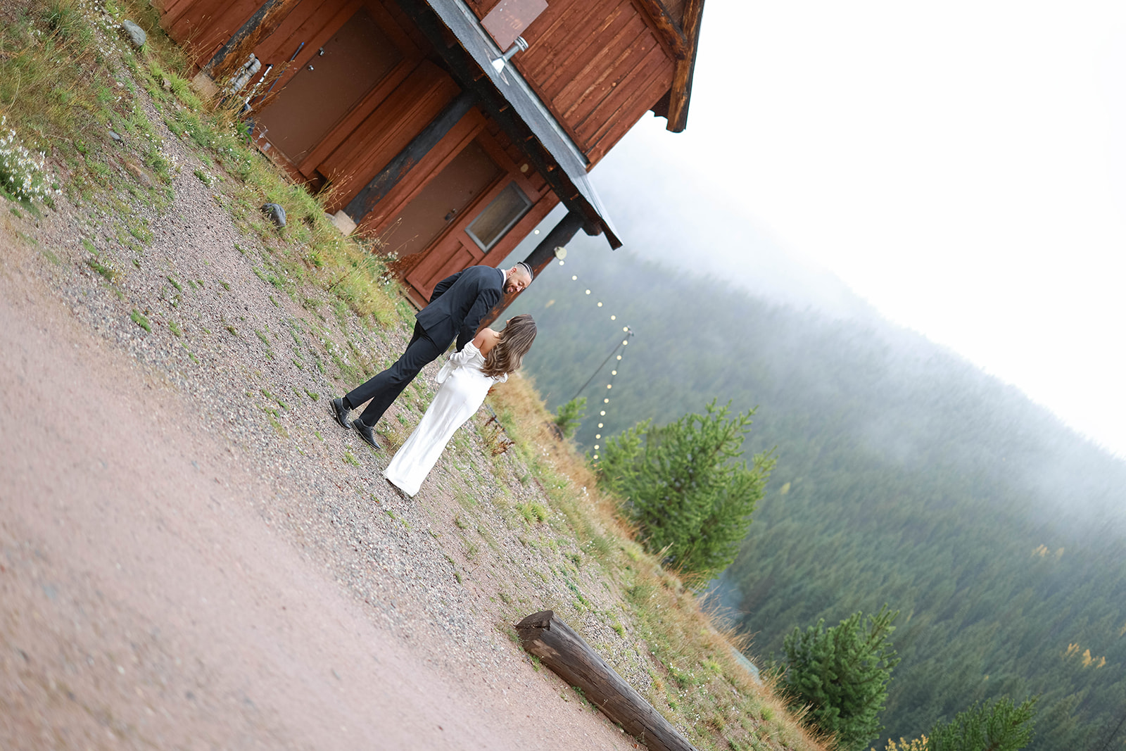 Bride and groom standing together outside a rustic cabin with string lights, surrounded by gravel paths and misty mountain views.