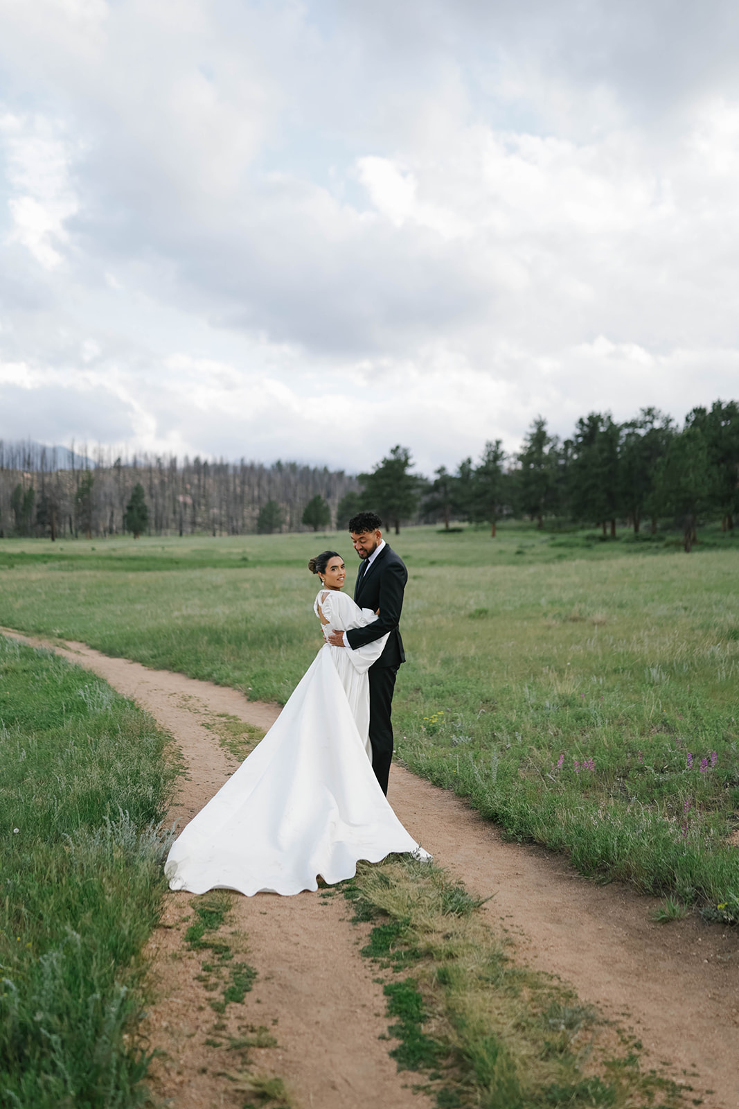 Bride and groom embracing on a quiet dirt path surrounded by open meadow and pine trees, her gown flowing behind her beneath a soft, cloudy sky.