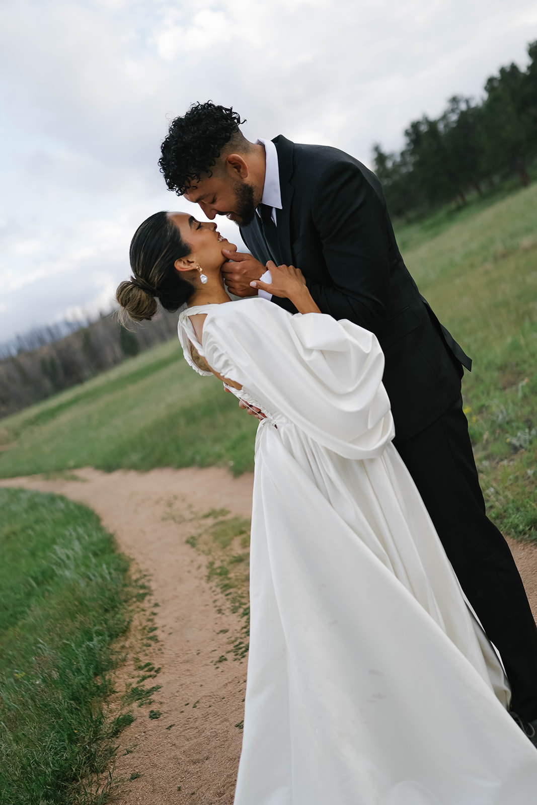 Romantic portrait of bride and groom standing along a winding trail in a mountain meadow, sharing a close embrace at sunset.