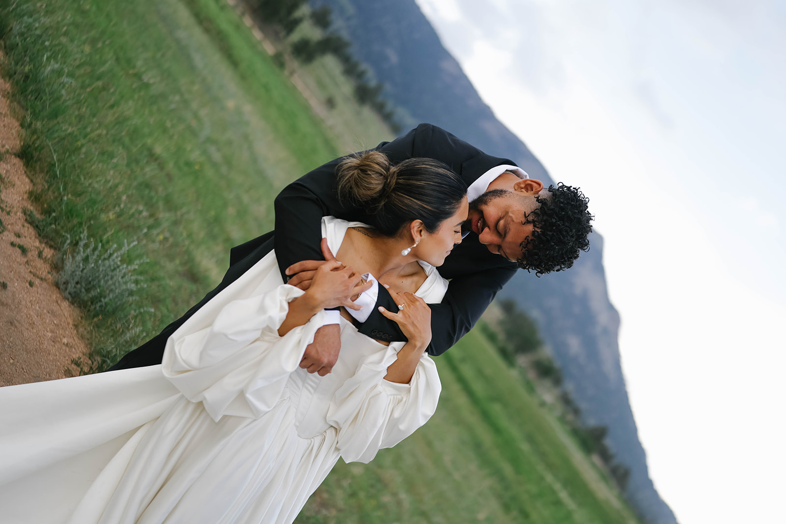 Bride and groom cuddle closely in a grassy field, her long train trailing across the earth during their Rocky Mountain National Park Elopement.