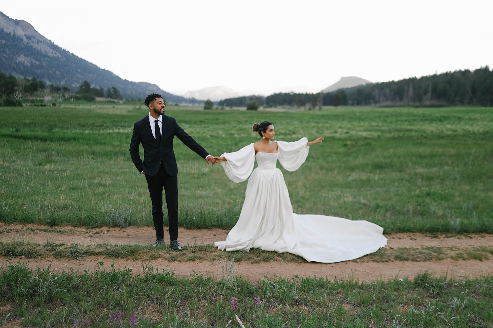 Bride and groom holding hands in an open meadow with mountain ridgelines behind them, her long-sleeve gown flowing dramatically during their Rocky Mountain National Park Elopement.