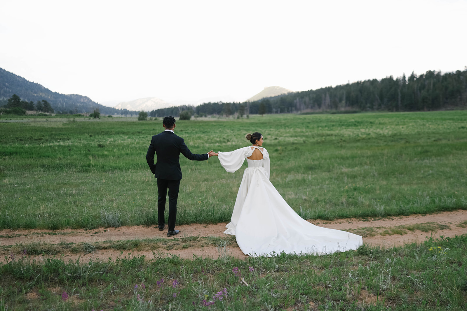 Bride and groom walking along a dirt trail through a wide green meadow with mountains in the background during a Rocky Mountain National Park Elopement.