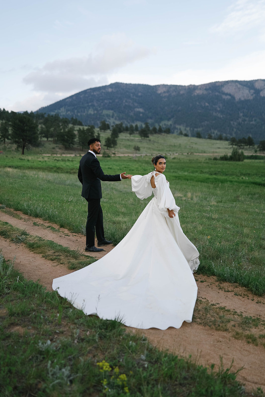 The bride glances back while holding hands with her groom along a dirt path in a wide open meadow.