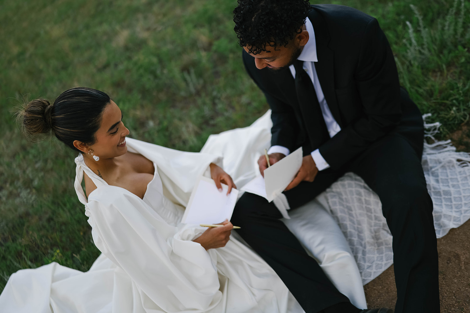 Overhead view of bride and groom smiling as they read personal vows to each other while seated on a blanket in an open Colorado field.