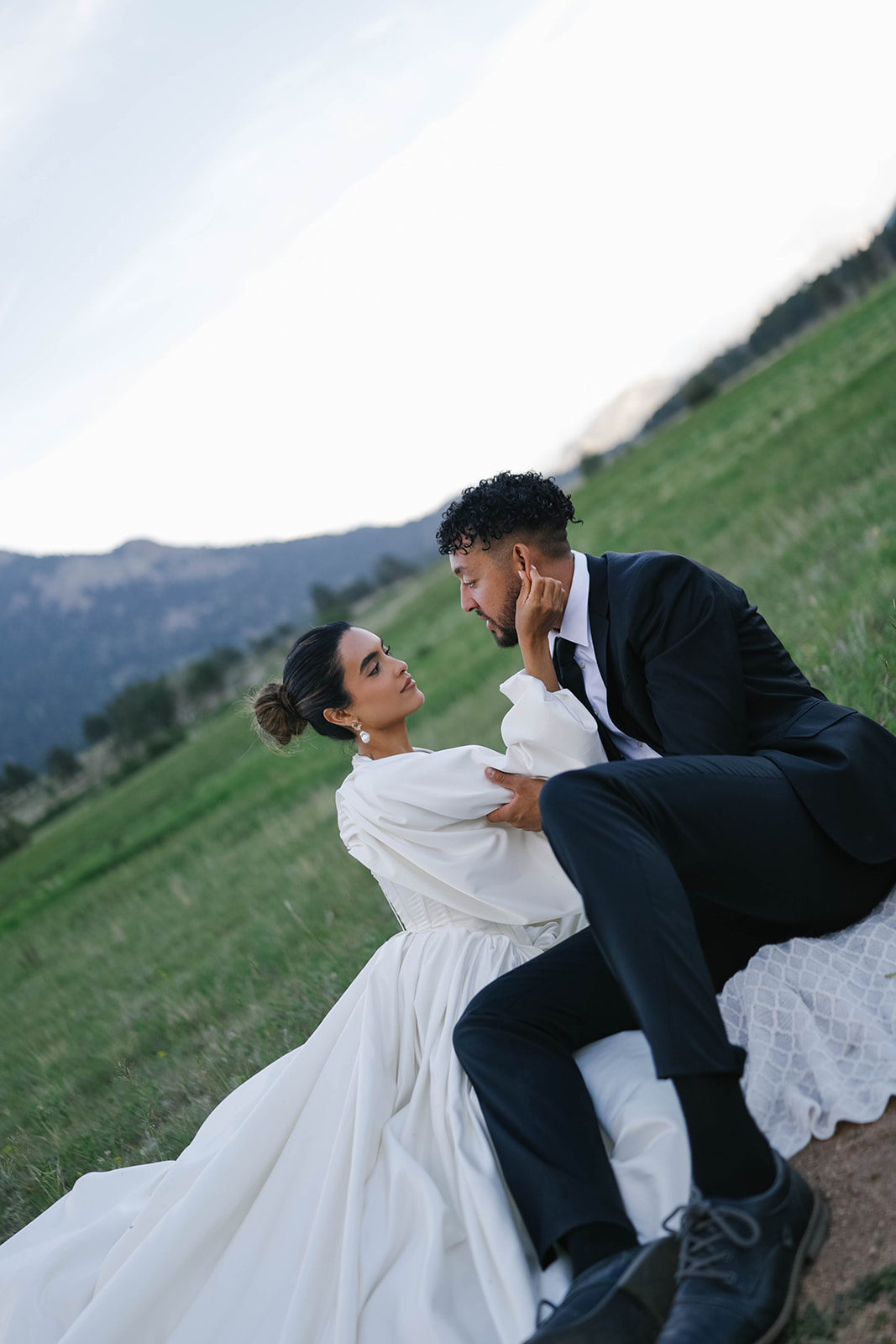 Bride and groom sitting close together on a blanket in a grassy field, sharing a soft, emotional moment.