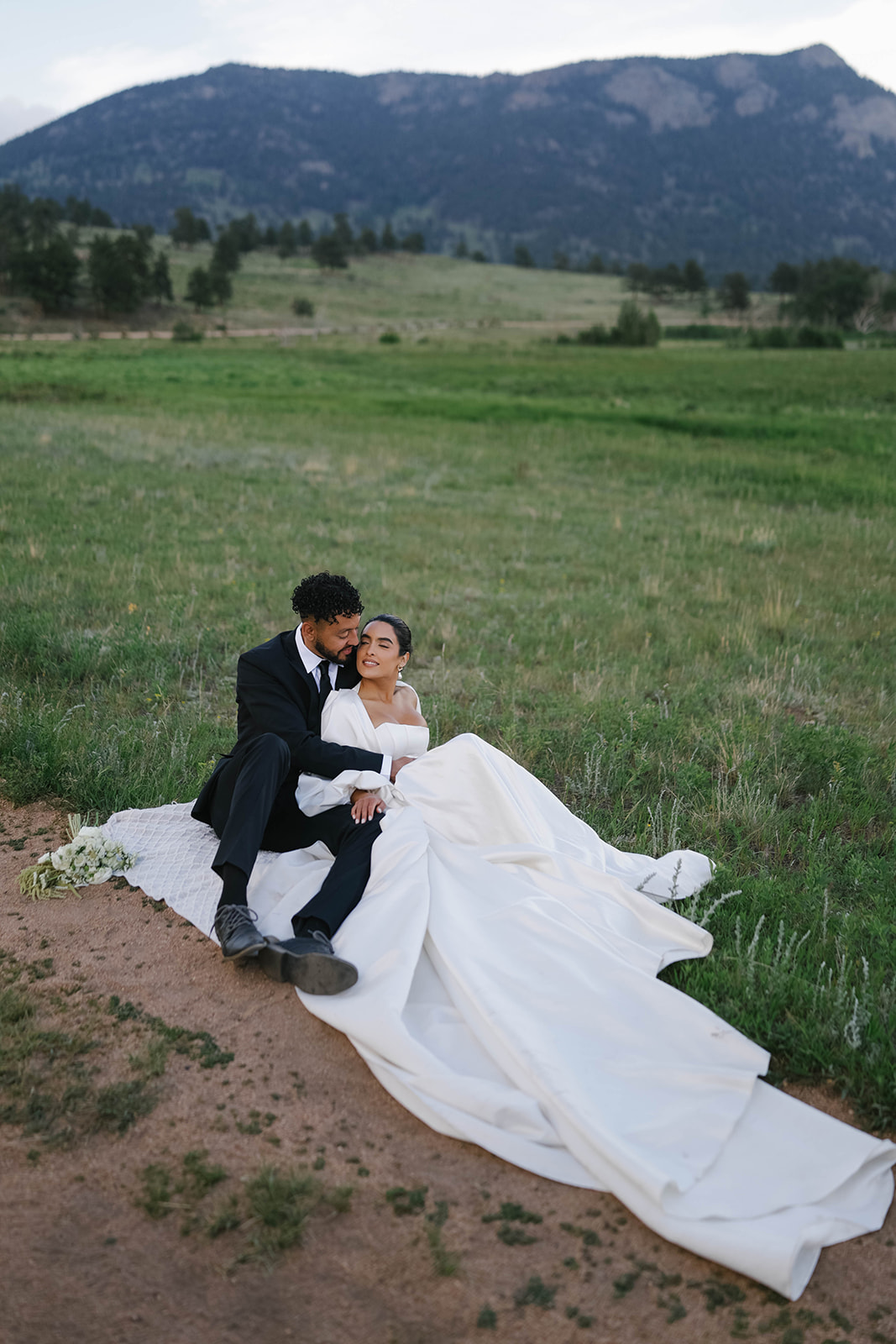Newlyweds sit together on a blanket in a meadow, bouquet nearby, sharing a quiet embrace during their Rocky Mountain National Park Elopement.