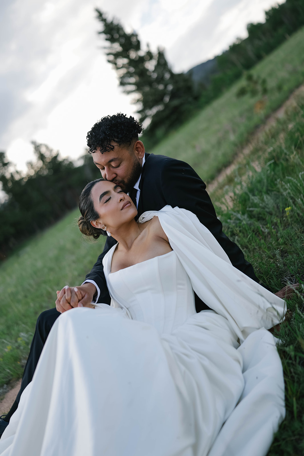 Intimate portrait of groom kissing the bride’s forehead as they sit together in a lush green meadow, her off-the-shoulder wedding dress softly draped around them.