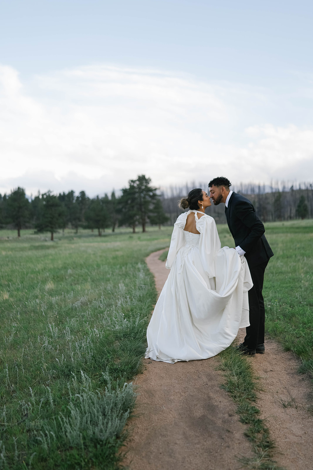 Bride and groom kissing on a winding dirt path surrounded by tall grass and pine trees in Rocky Mountain National Park.