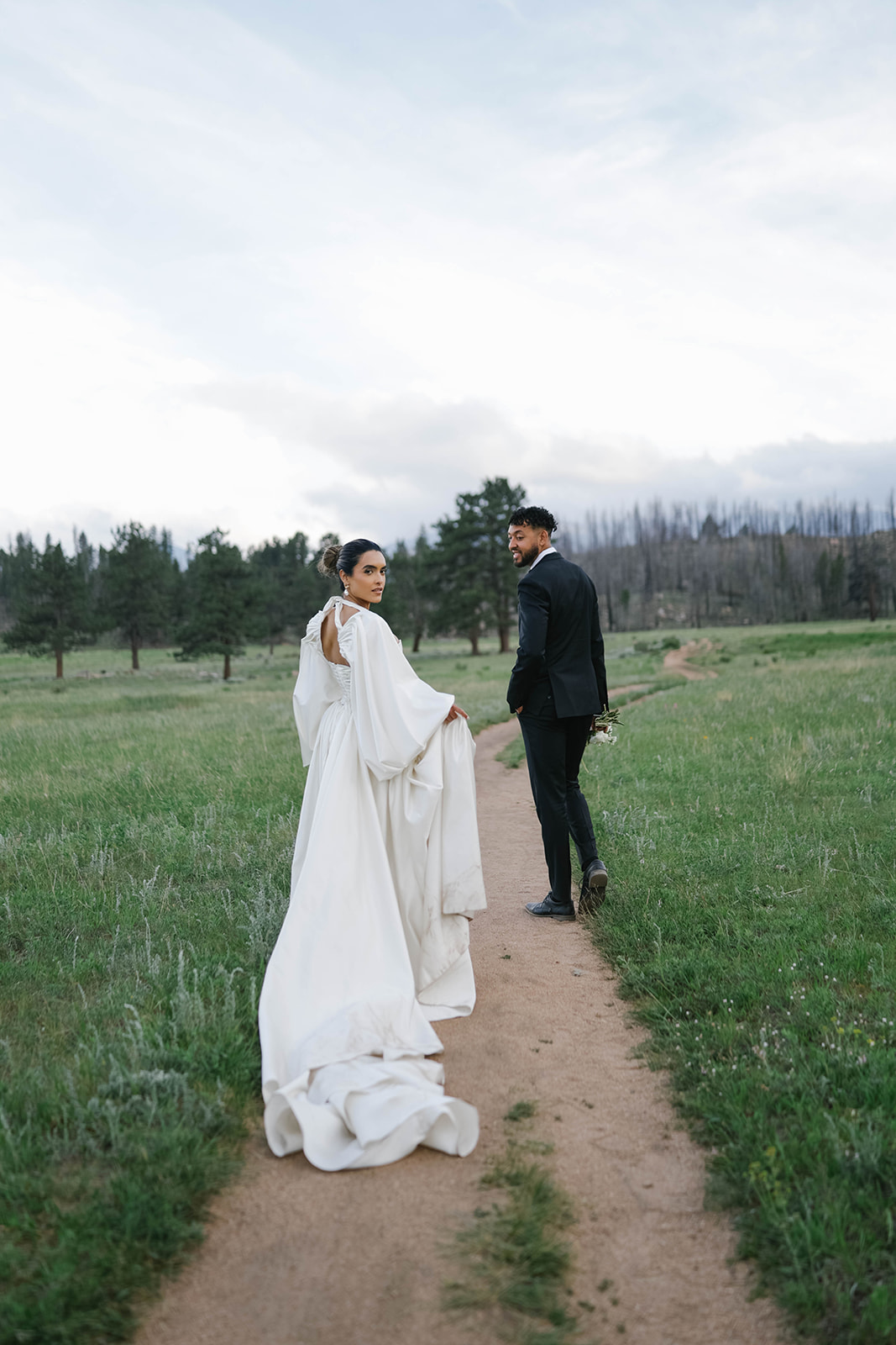 Bride and groom walking hand-in-hand down a dirt path through a green meadow, her long ivory train flowing behind her during their Rocky Mountain National Park Elopement.