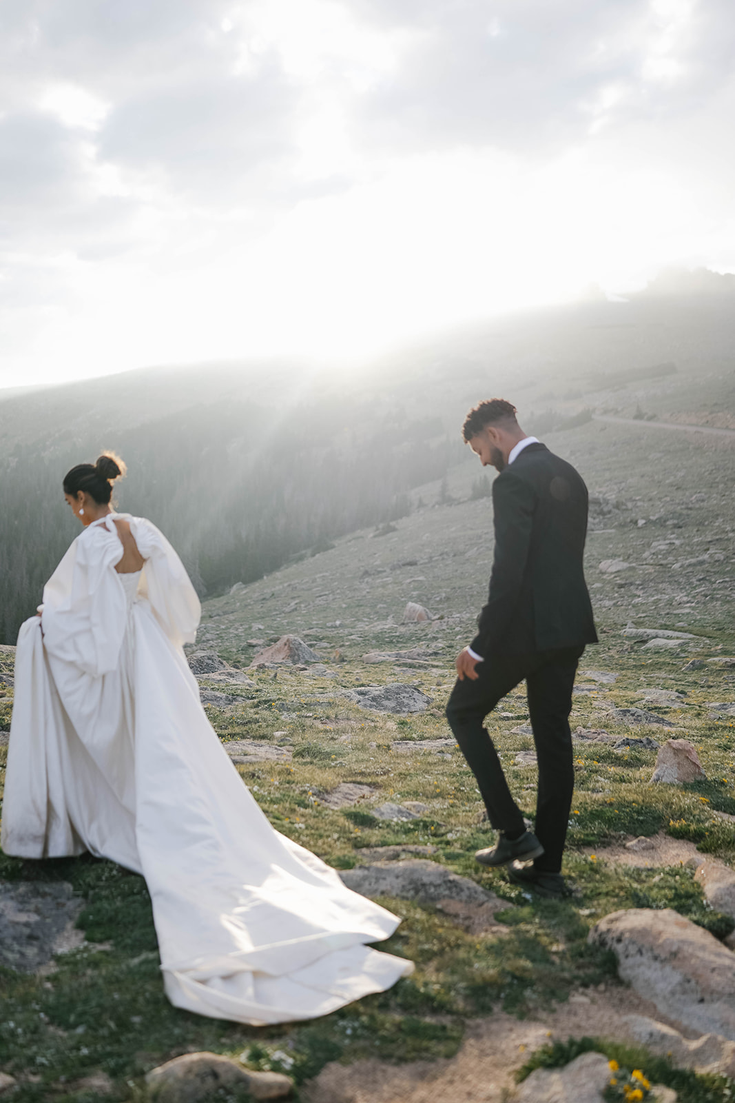 Newly married couple walking across a sunlit alpine ridge with dramatic mountain scenery during their Rocky Mountain National Park Elopement.