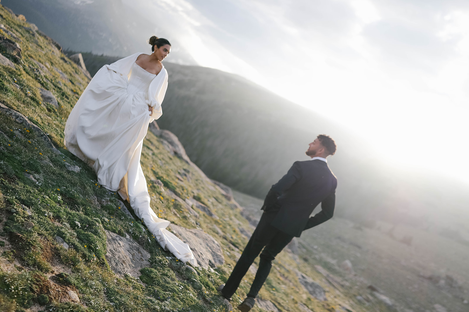 Bride standing on a mountainside at golden hour as the groom approaches, sunlight glowing behind them during their Rocky Mountain National Park Elopement in the high alpine.