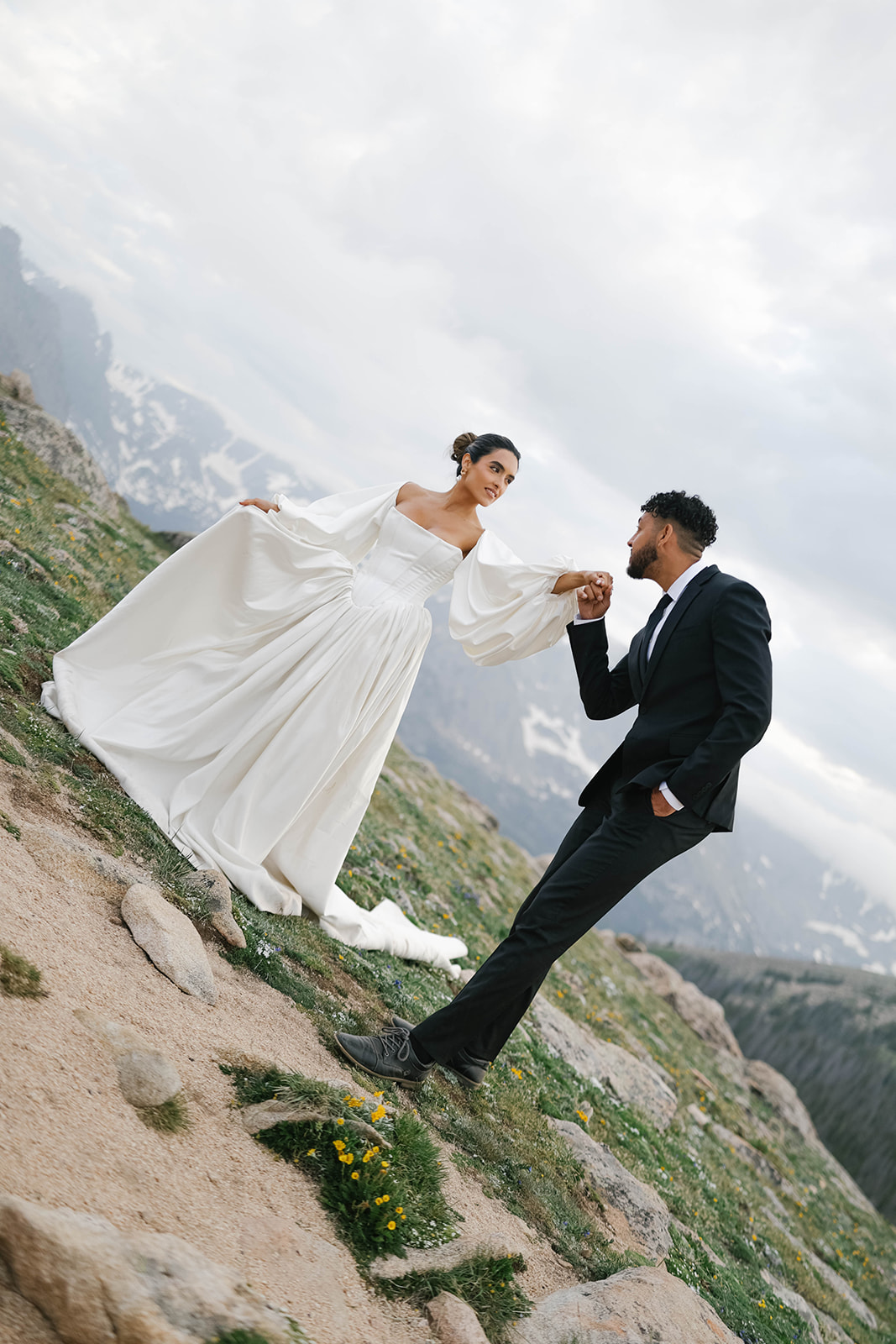 Bride and groom holding hands on a sloped alpine trail with dramatic mountain views during their Rocky Mountain National Park Elopement, her flowing long-sleeve gown catching the wind as he leans in.
