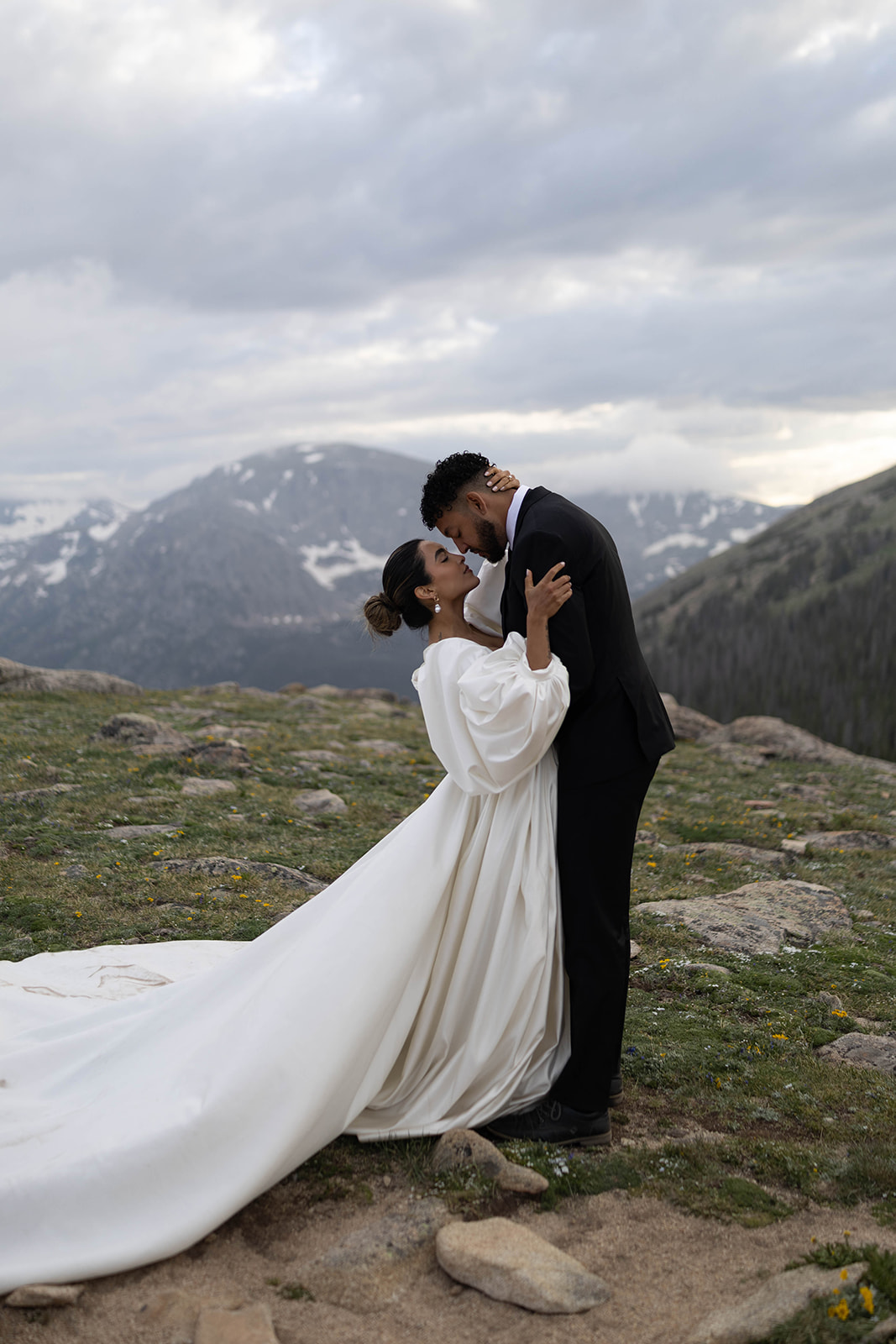 Intimate portrait of a bride and groom embracing on a mountain overlook, her long-sleeved gown flowing against a dramatic Rocky Mountain National Park Elopement backdrop.