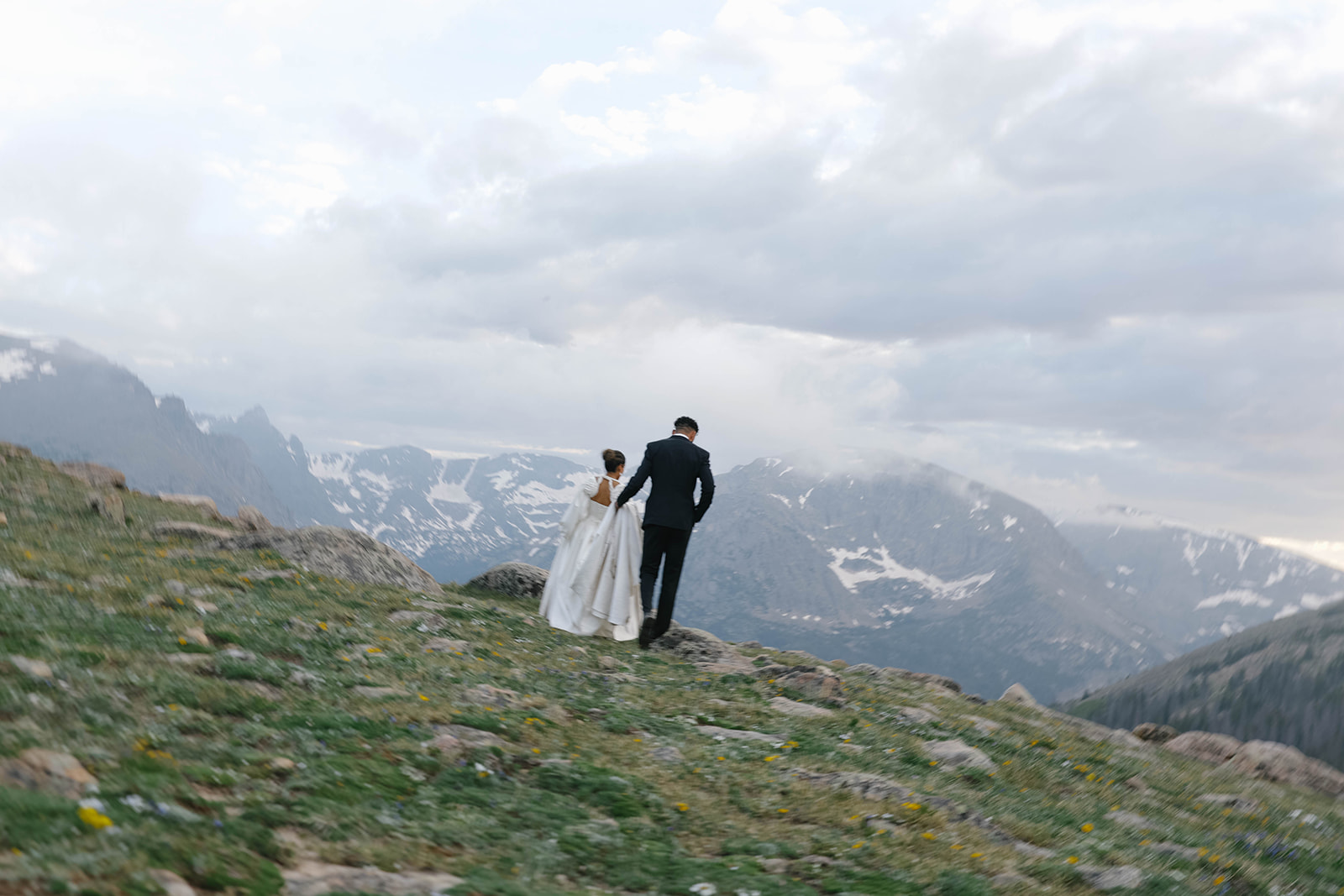 Bride and groom walk hand in hand along a rocky alpine ridge during their Rocky Mountain National Park Elopement, with dramatic snow-capped peaks stretching across the horizon.