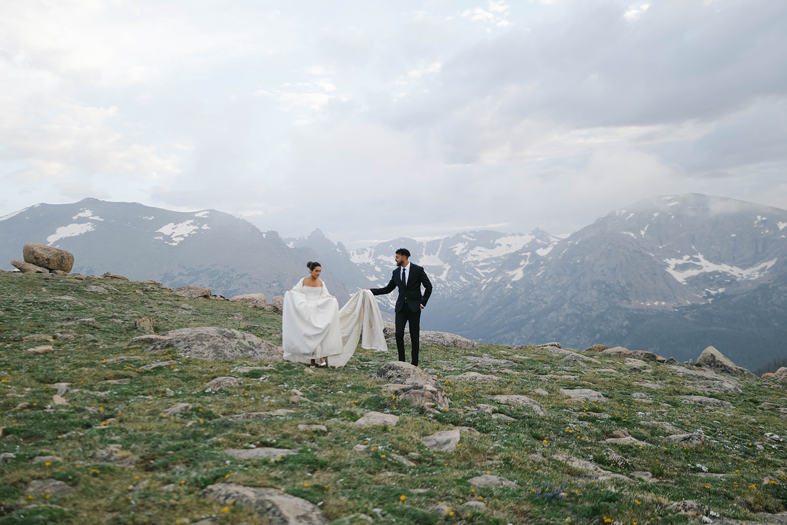 Bride and groom walk across an alpine ridge with snow-capped peaks behind them during their Rocky Mountain National Park Elopement.