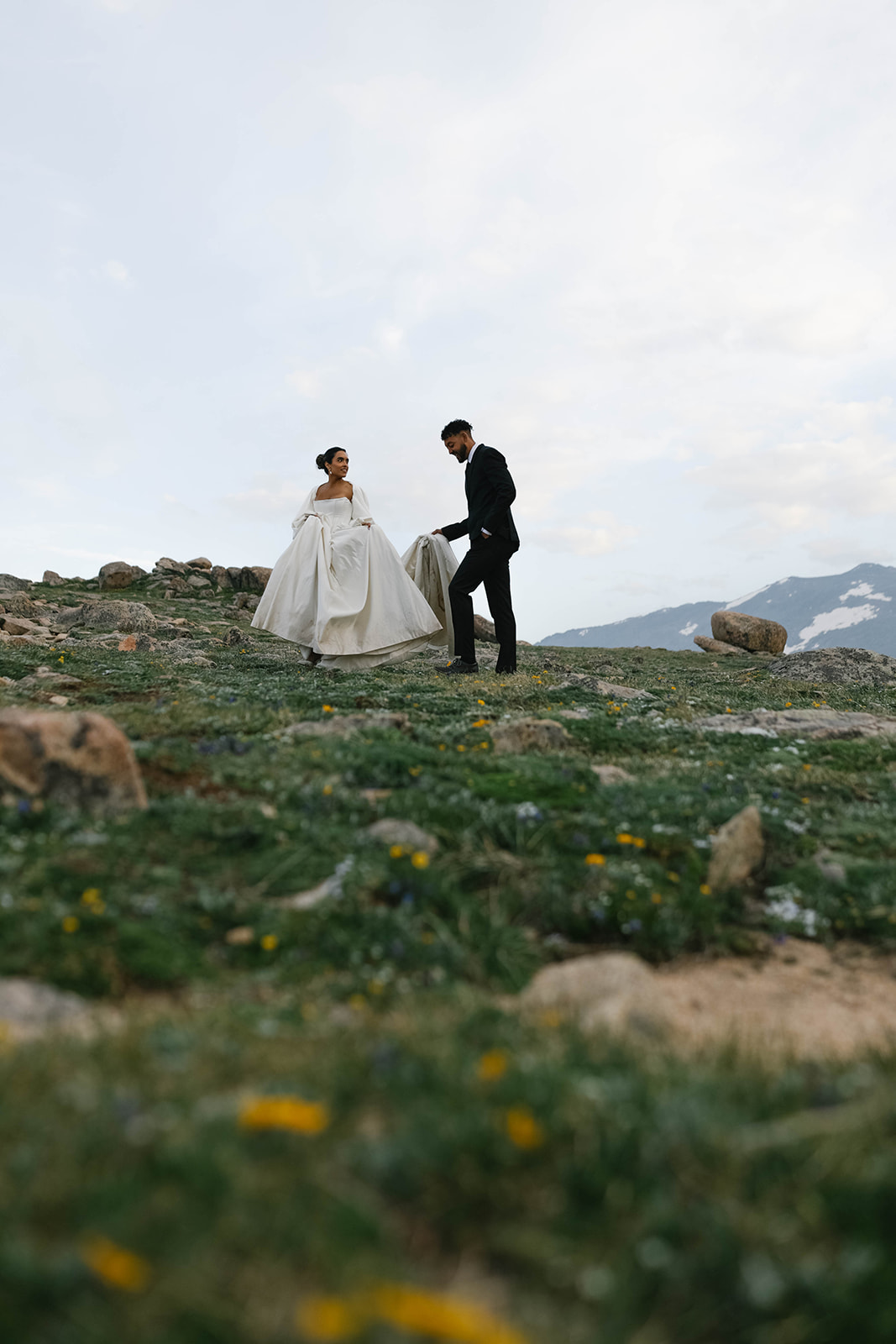 Wide landscape shot of bride and groom walking hand in hand across a rocky alpine field with expansive skies and mountain peaks in the distance at Rocky Mountain National Park.