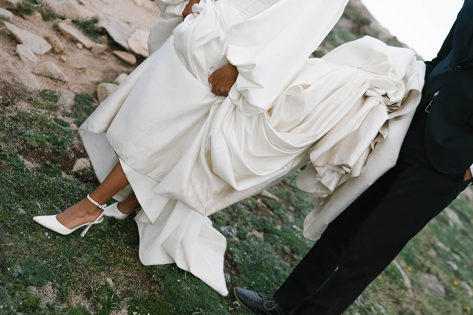 Close-up detail of bride lifting her gown to reveal white heels as she walks across rocky alpine terrain during her Rocky Mountain National Park Elopement.
