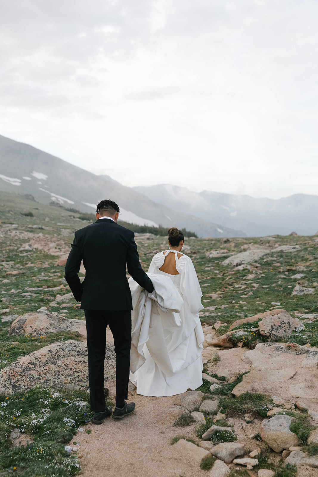 Couple seen from behind hiking across rocky terrain at sunset, the bride lifting her flowing gown as they explore the mountains during their Rocky Mountain National Park Elopement.