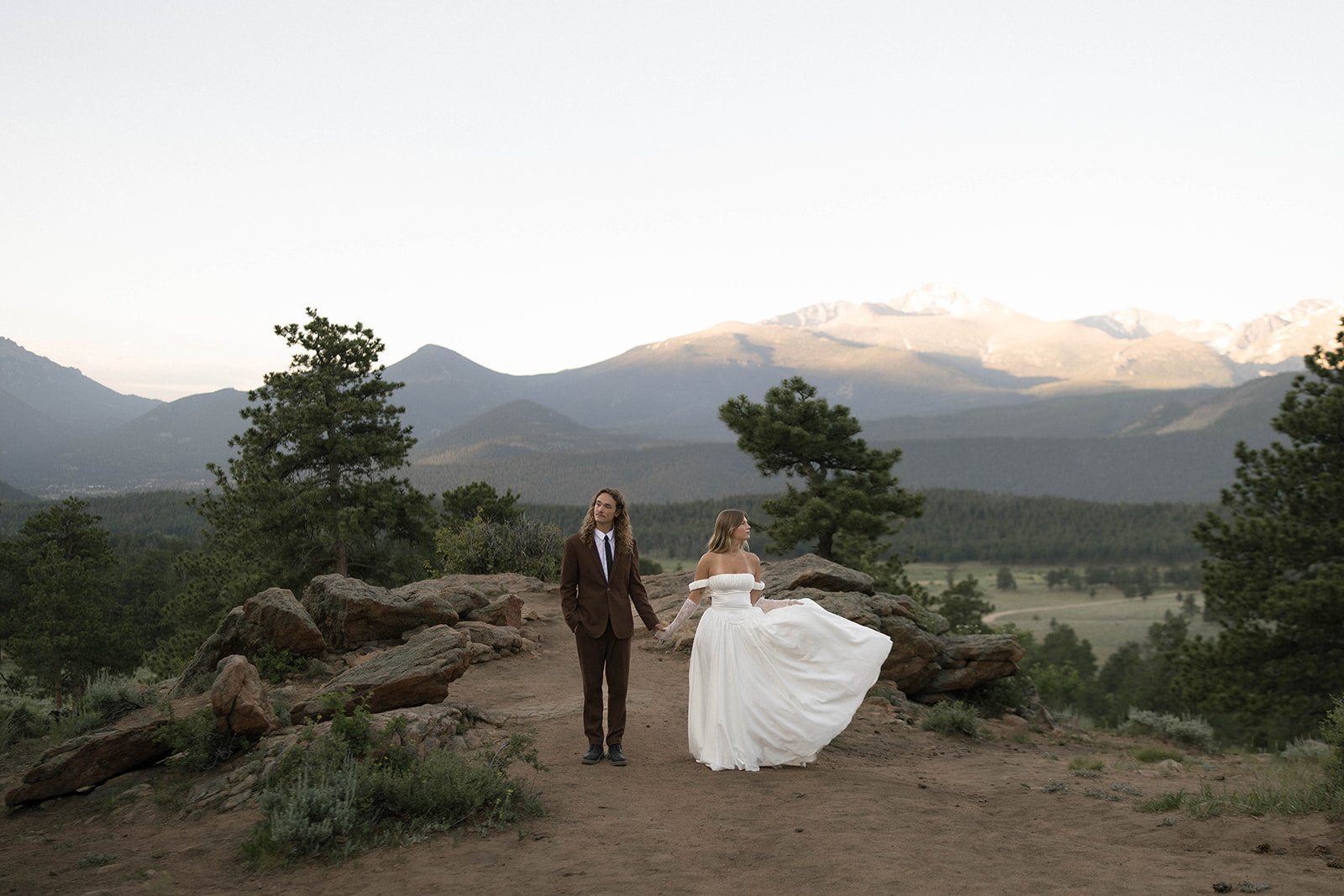 Couple stands hand in hand on a scenic cliffside with dramatic mountain peaks in the background, capturing the beauty of their Rocky Mountain National Park elopement at golden hour.