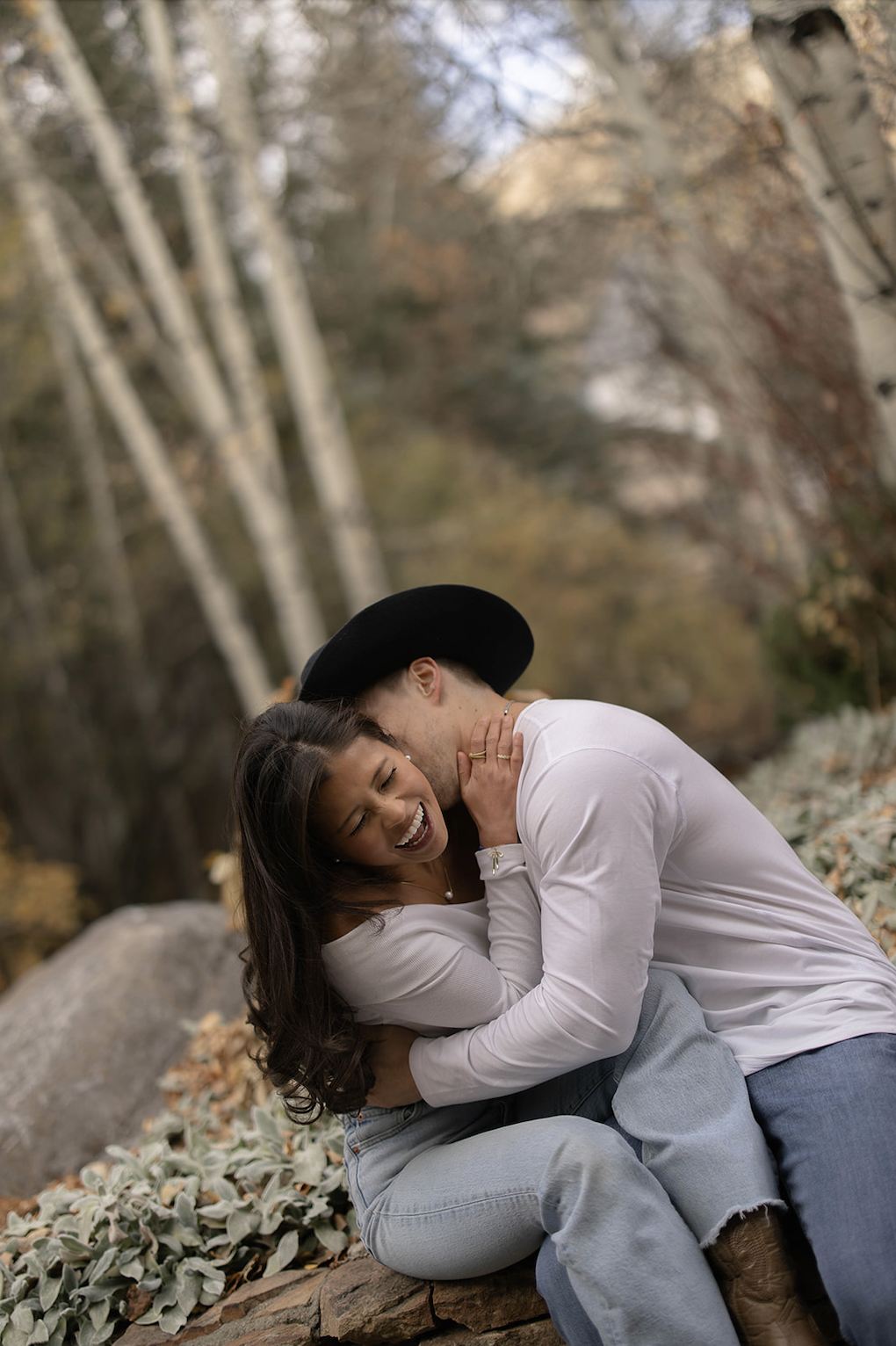 Playful couple laughing and cuddling on a stone wall beneath tall aspens during fall in the mountains near Vail, Colorado.