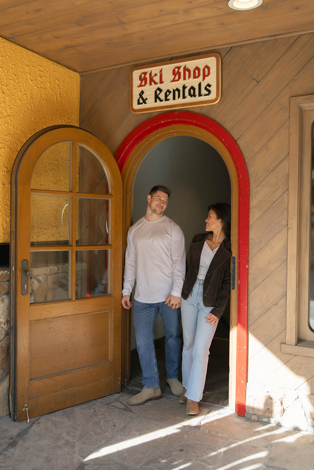 Couple holding hands as they step out of a charming ski shop doorway in Vail Village, Colorado, surrounded by warm wood tones and alpine architecture.